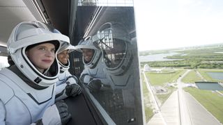 Two women in spacesuits looking out a window showing a landscape of trees, water and roads