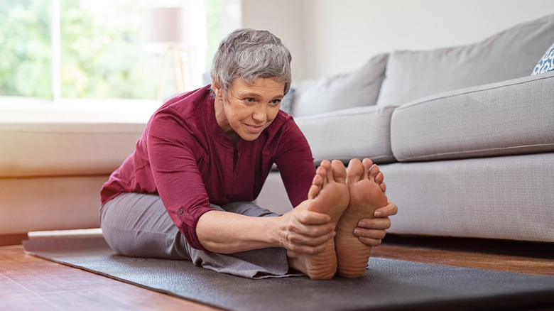 An elderly woman is sitting on the exercise mat holding her toes